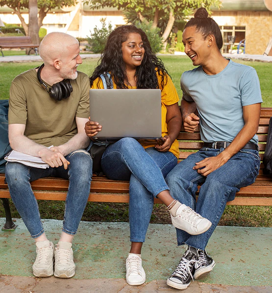 Three students sitting on a park bench, laughing around a laptop